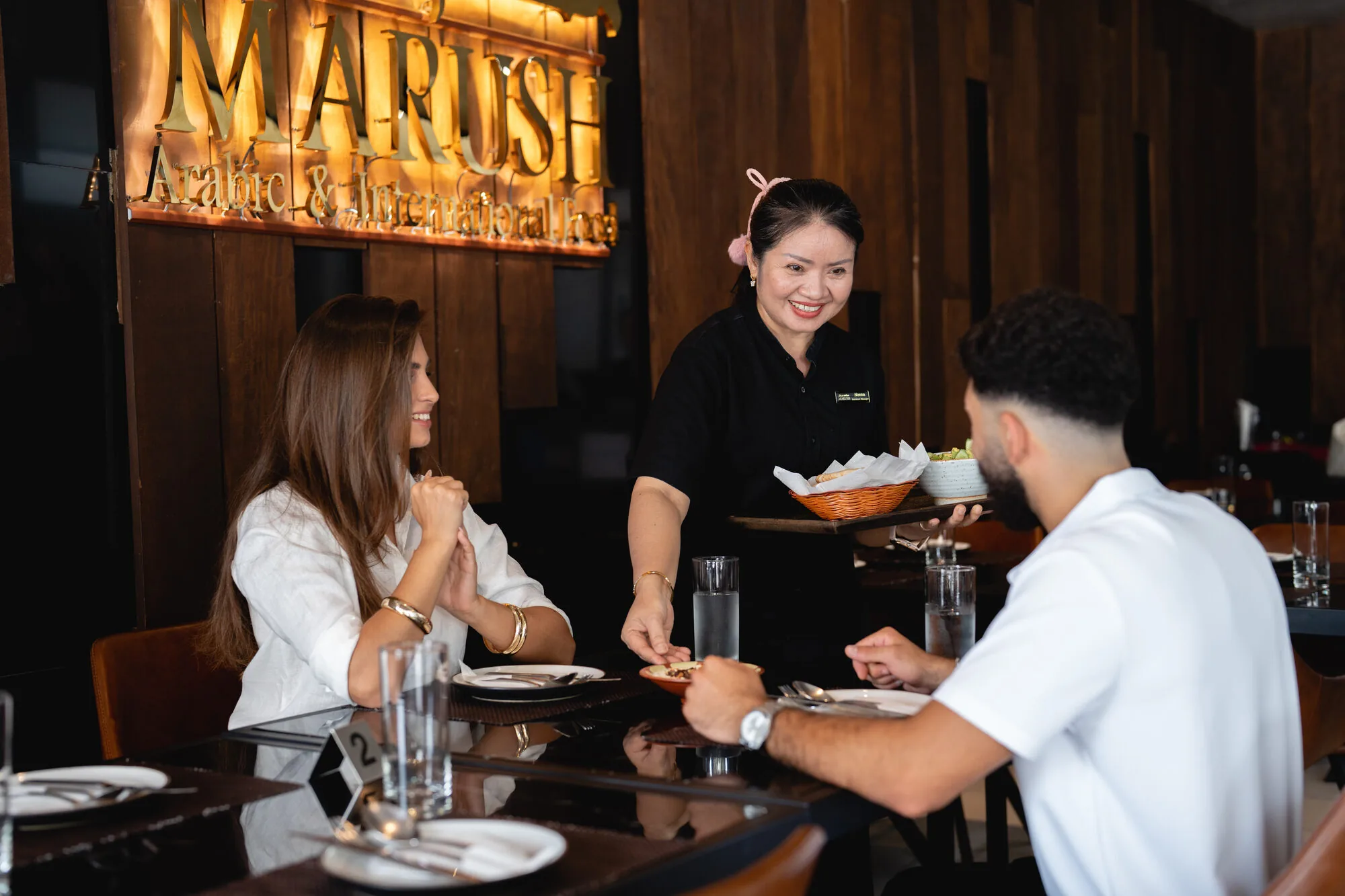 Marush waitress serving dishes to a smiling couple.